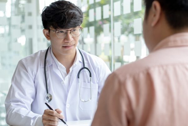 Doctor in a white coat with a stethoscope consulting a male patient across a desk in a bright clinic setting.