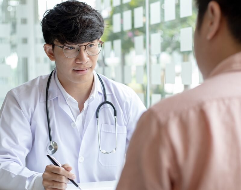 Doctor in a white coat with a stethoscope consulting a male patient across a desk in a bright clinic setting.