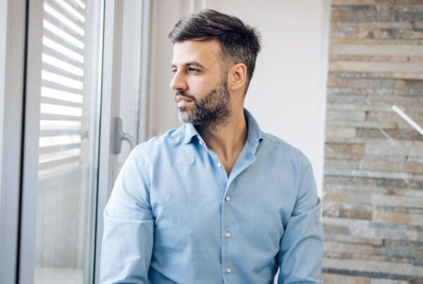Man in a blue shirt looking out a window in a bright room.